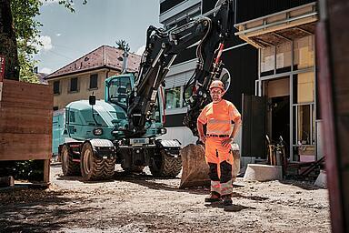 Operator wearing high-visibility workwear standing beside a Wacker Neuson EW100 wheeled excavator at an urban construction site.