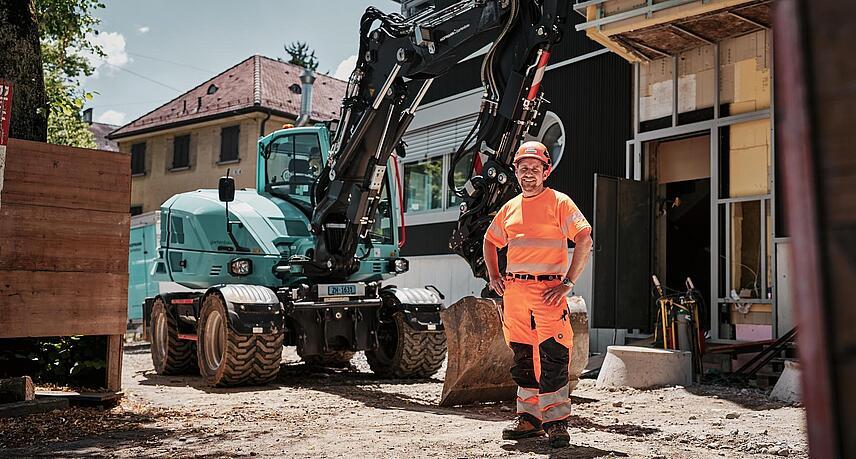 Operatore che indossa indumenti da lavoro ad alta visibilità, in piedi accanto a un escavatore gommato Wacker Neuson EW100 in un cantiere urbano.