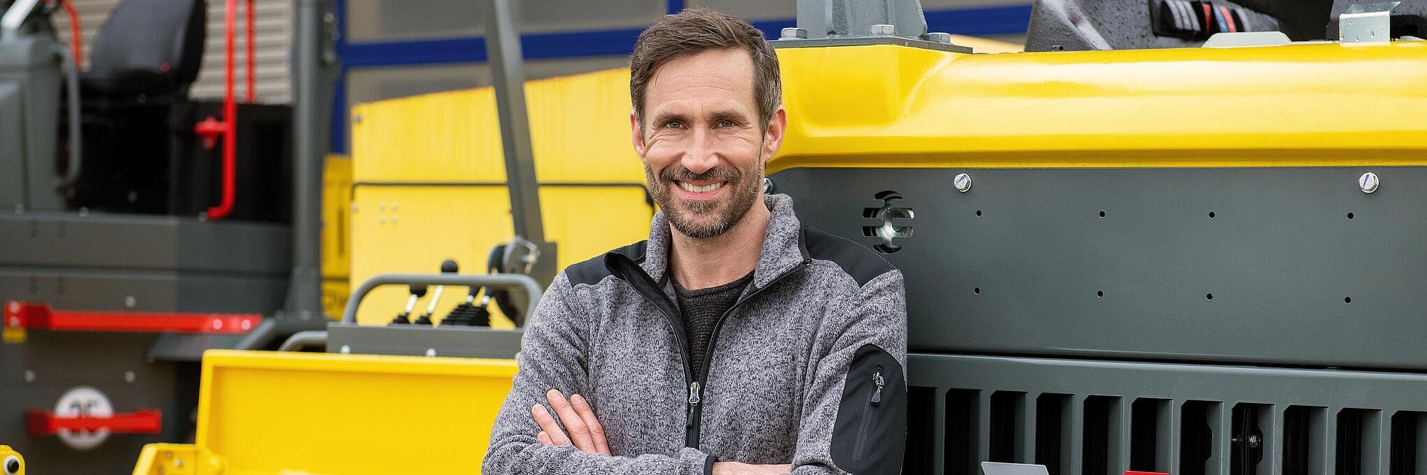 Una sorridente dipendente Wacker Neuson si appoggia a una macchina edile Wacker Neuson con le braccia piegate.