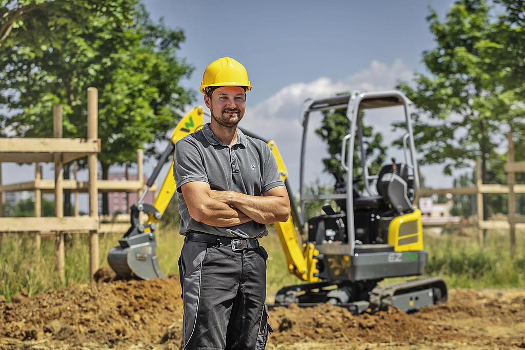 Ein Mann mit gelbem Schutzhelm steht vor einem Wacker-Neuson-Bagger auf einer Baustelle.