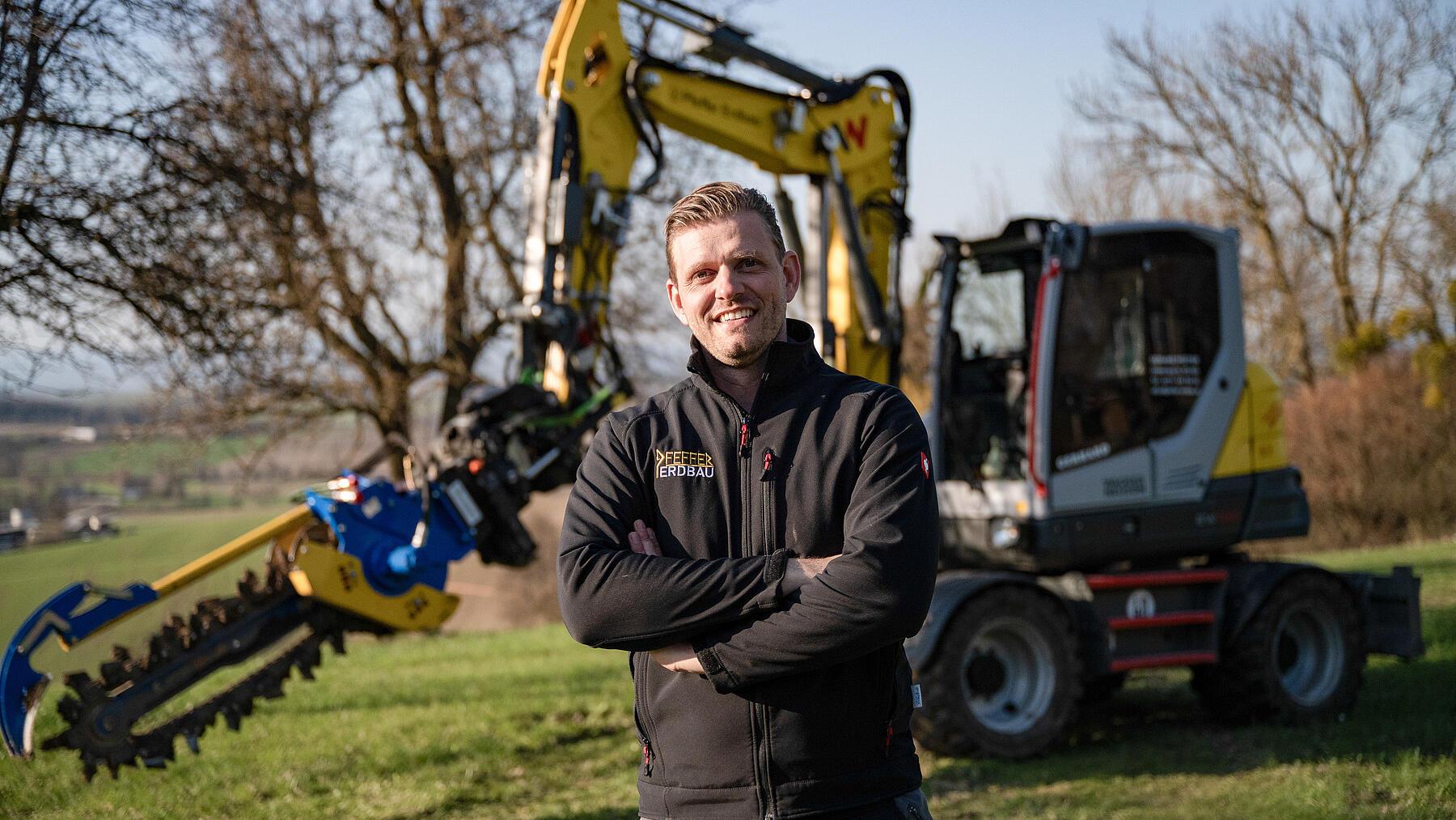 Ein Bauunternehmer steht vor einem Mobilbagger vom Typ Wacker Neuson EW100, der mit einem Grabenfräsaufsatz ausgestattet ist, in einem Arbeitsbereich in ländlicher Umgebung.
