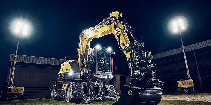 Wacker Neuson EW135 wheeled excavator equipped with a tiltrotator and grading bucket, illuminated under floodlights in a nighttime work environment.