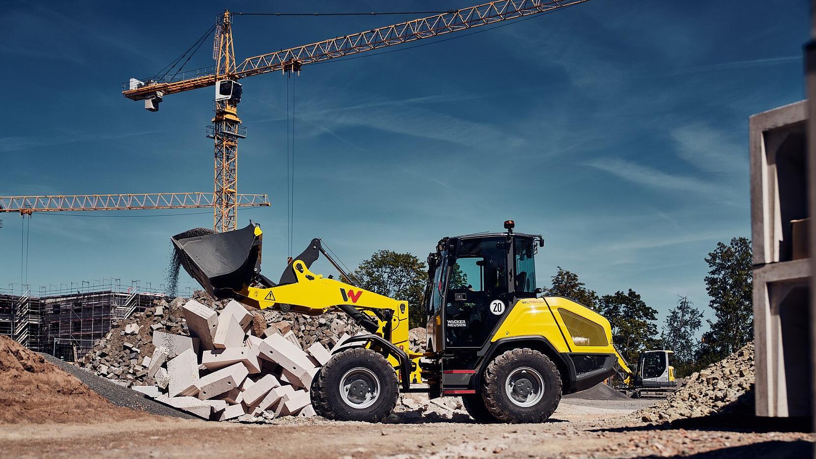 Wacker Neuson WL950p wheel loader unloading rubble from a bucket onto a demolition debris pile at a construction site with tower cranes in the background.