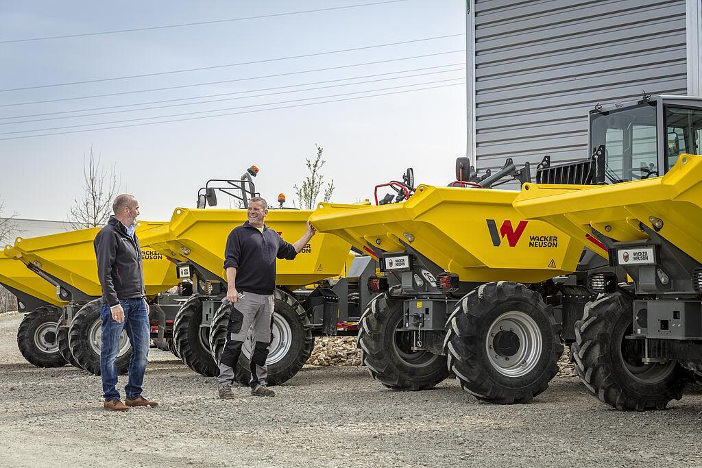 Two men are standing next to several yellow Wacker Neuson dumpers outdoors.
