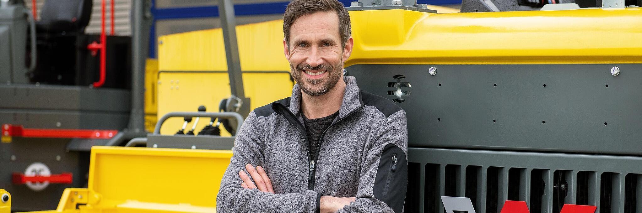 Lächelnder Wacker Neuson Mitarbeiter lehnt mit verschränkten Armen gegen eine Wacker Neuson Baumaschine.